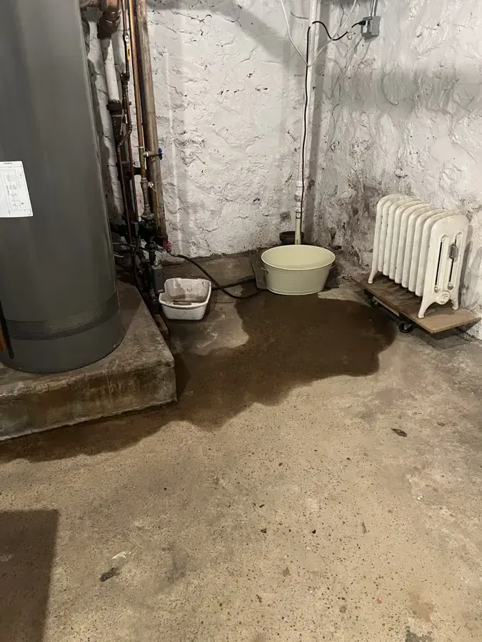 An old whitewashed stone basement with a cast iron radiator and water on the floor, the kind of space that floods in Brookline homes
