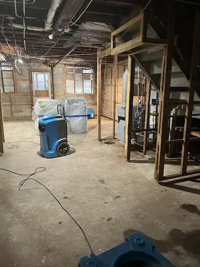 A dehumidifier and an air mover working in a basement opened to the framing, part of a water damage dry-out in Milton, MA