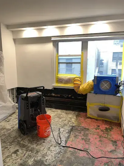 Dehumidifier and air mover drying a Dedham room stripped of its flooring, with an orange bucket on the bare subfloor
