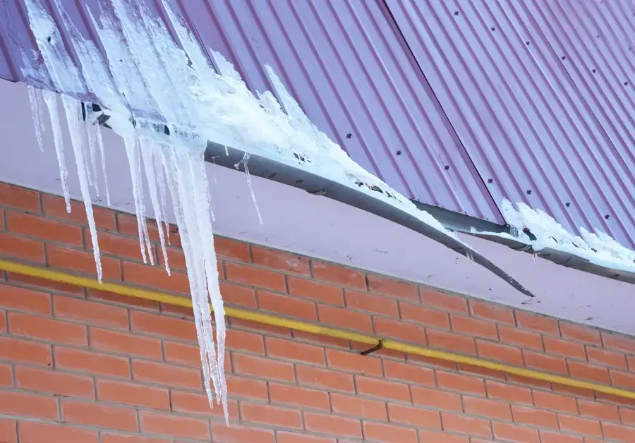 Icicles and a ridge of ice along a roof edge above a brick wall, ice dam roof leak water damage in Brookline, MA